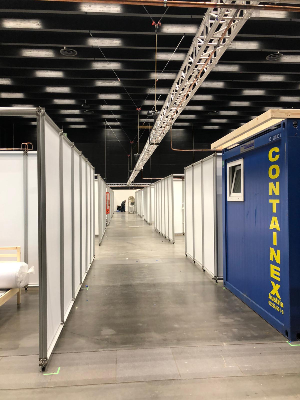 Innenansicht der Messehalle mit langen Reihen modularer Trennwände, die einen Gang zu temporären Raum- bzw. Klinikbereichen bilden; rechts ein blauer Container.